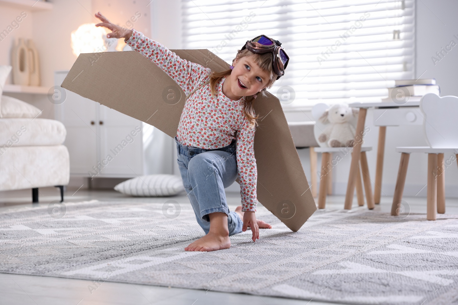 Photo of Cute little girl with cardboard plane wings and goggles playing pilot at home