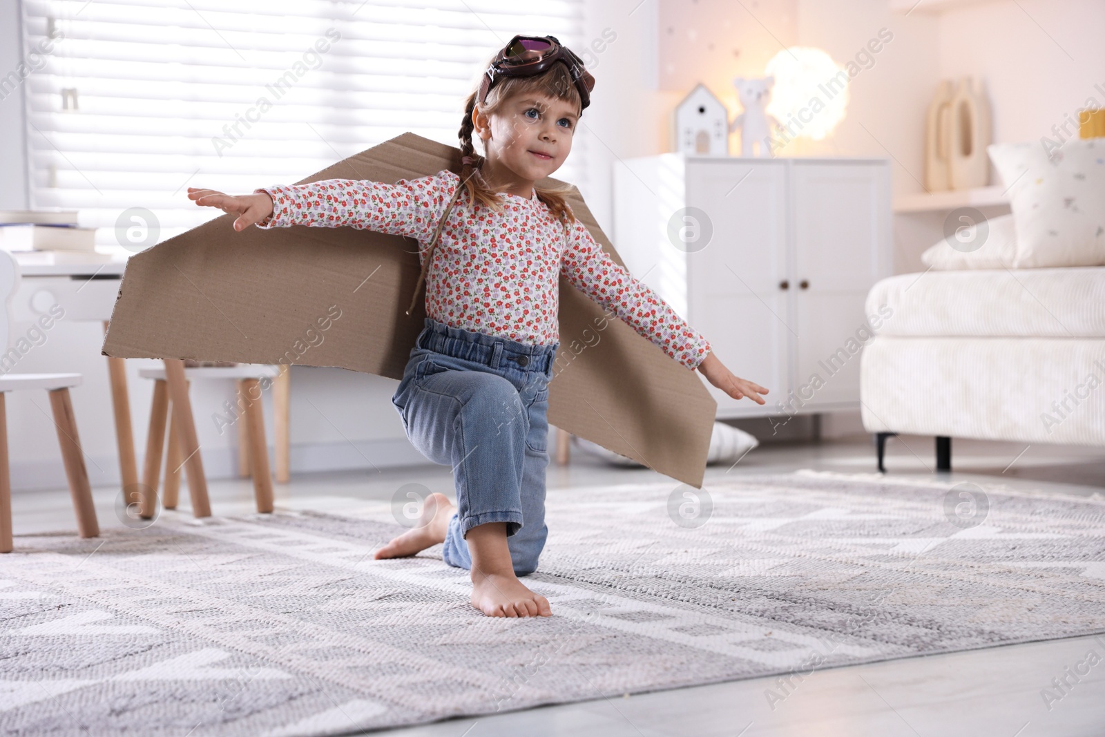 Photo of Cute little girl with cardboard plane wings and goggles playing pilot at home