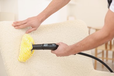 Photo of Professional janitor steam cleaning armchair in room, closeup