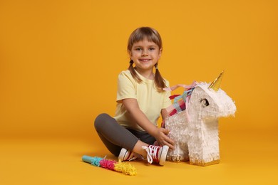 Photo of Happy girl with unicorn shaped pinata and stick on orange background