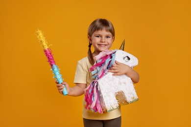 Photo of Happy girl with unicorn shaped pinata and stick on orange background