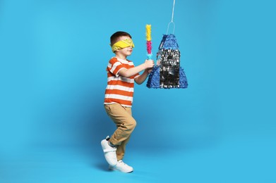 Photo of Cute boy with tied eyes breaking rocket shaped pinata on light blue background