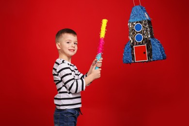 Photo of Happy boy breaking rocket shaped pinata with stick on red background