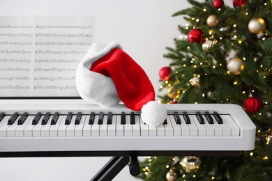 Photo of Synthesizer with music sheets and Santa hat near Christmas tree indoors