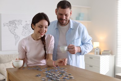 Photo of Happy couple with cups of coffee solving puzzle together at wooden table indoors