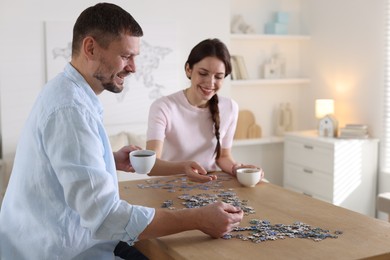 Photo of Happy couple with cups of coffee solving puzzle together at wooden table indoors