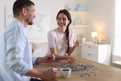 Photo of Happy couple with cups of coffee solving puzzle together at wooden table indoors