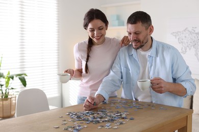 Photo of Happy couple with cups of coffee solving puzzle together at wooden table indoors