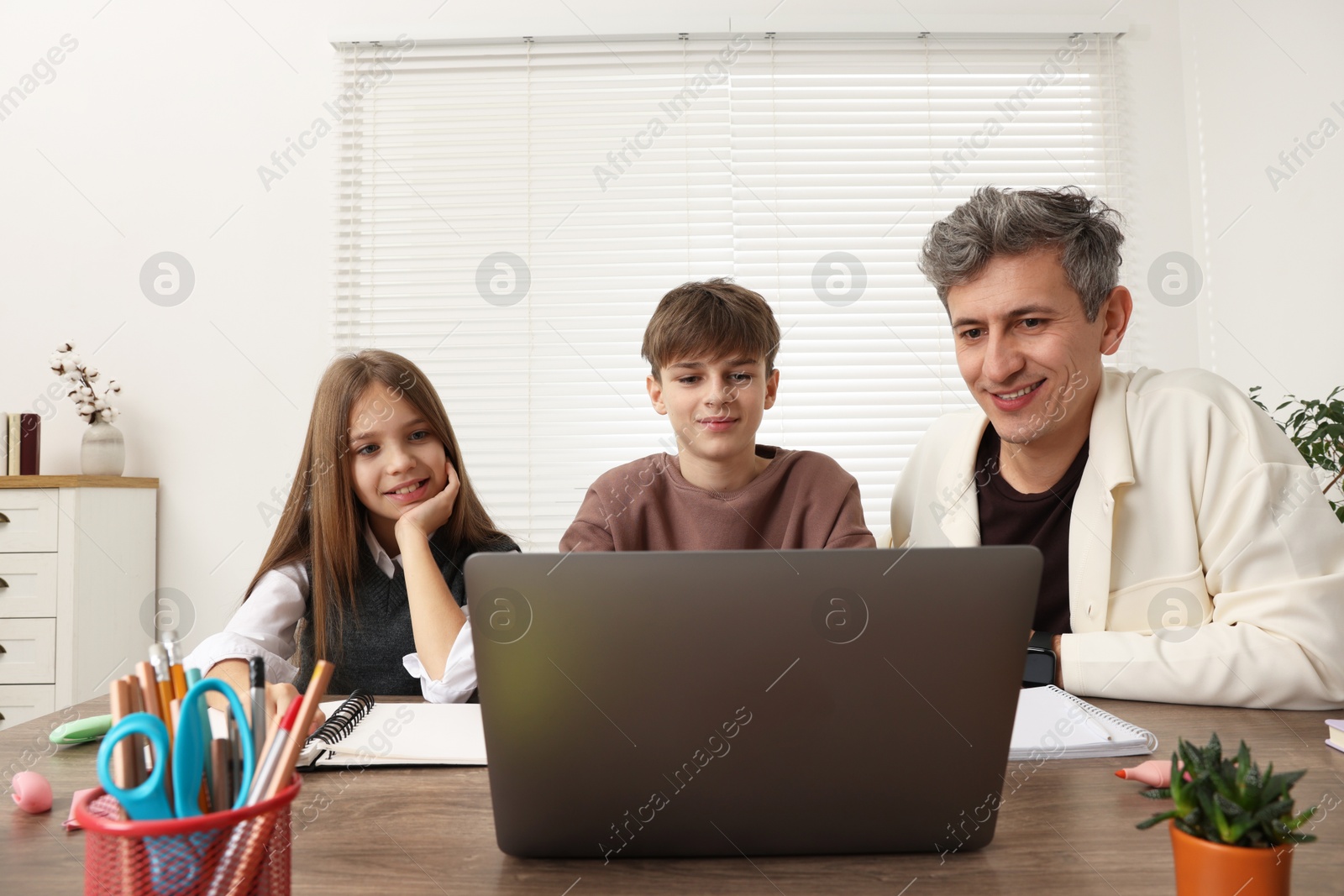 Photo of Smiling father and his children doing homework with laptop at table indoors