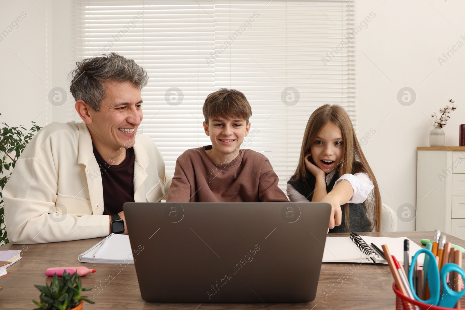 Photo of Smiling father and his children doing homework with laptop at table indoors