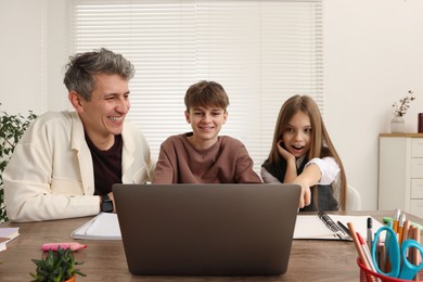 Photo of Smiling father and his children doing homework with laptop at table indoors