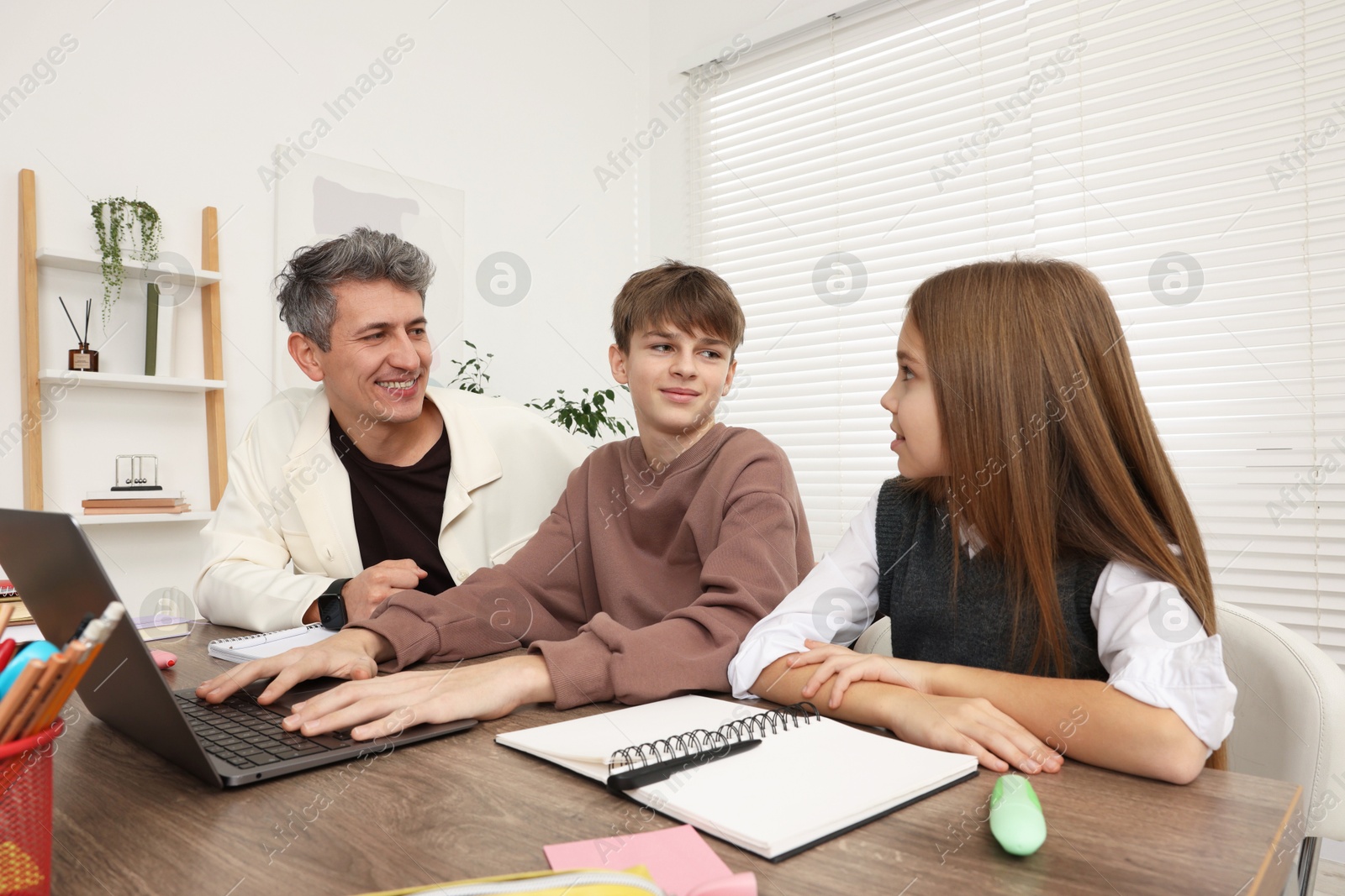 Photo of Smiling father and his children doing homework with laptop at table indoors