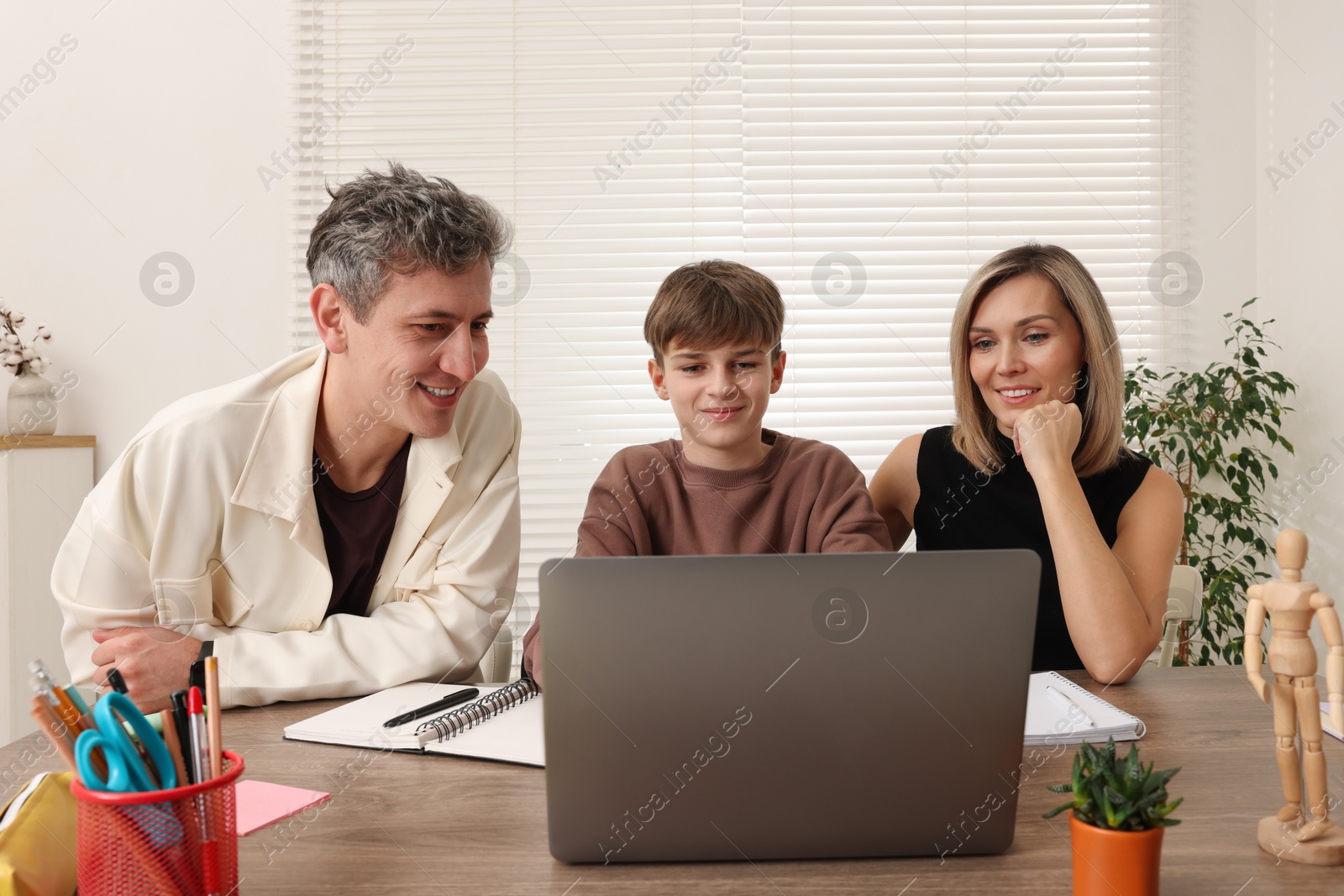 Photo of Smiling parents and their son doing homework with laptop at table indoors