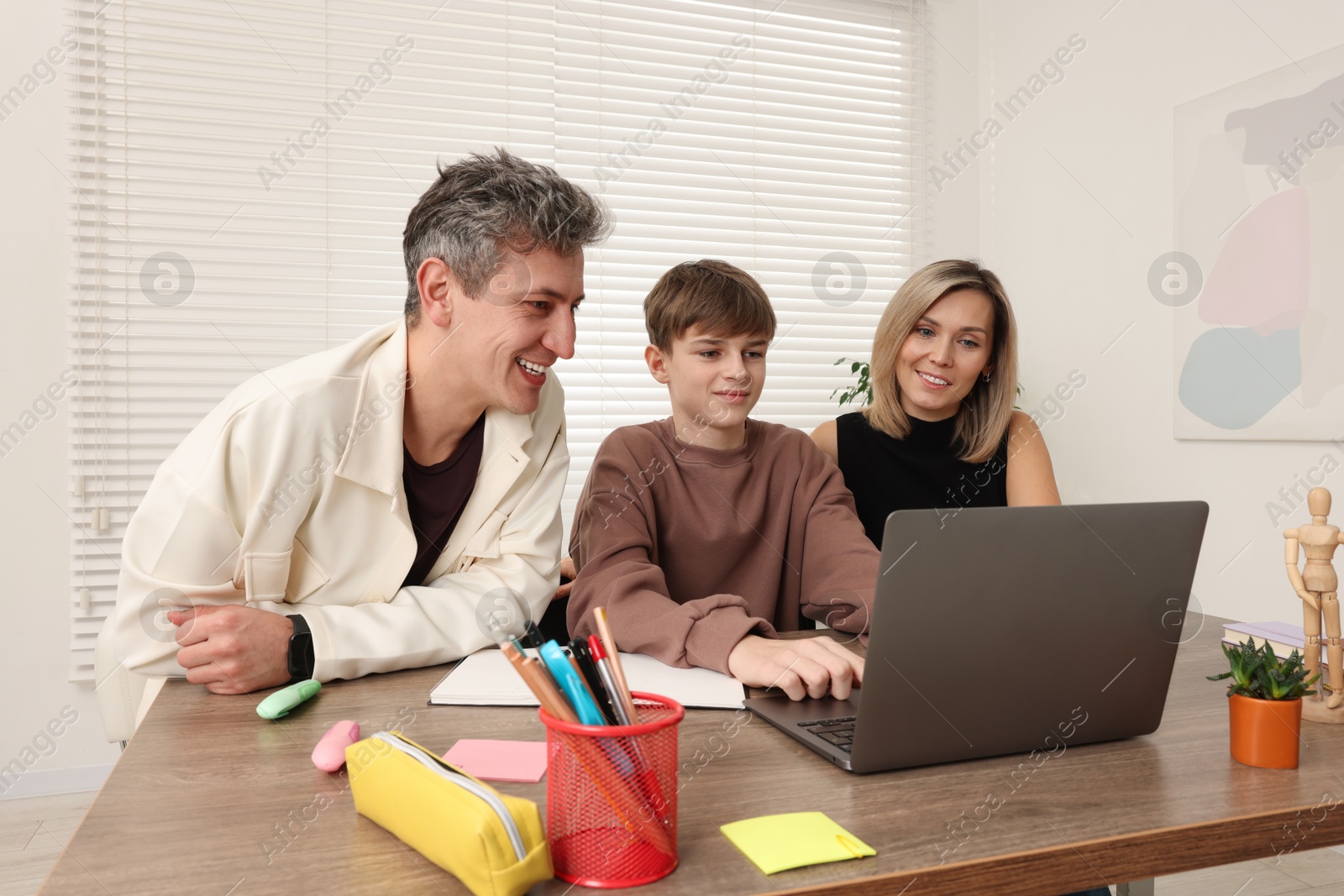 Photo of Smiling parents and their son doing homework with laptop at table indoors