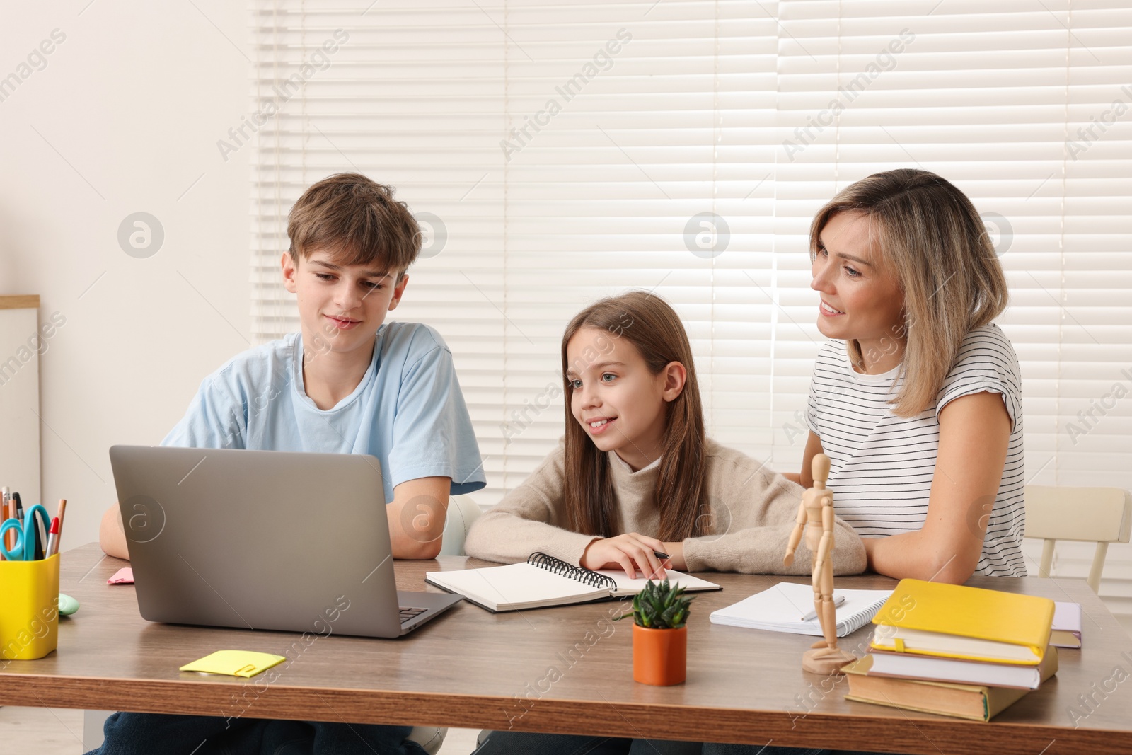 Photo of Smiling mother helping her children with homework at table indoors