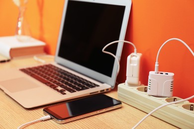 Photo of USB adapters with cables plugged into power strip charging laptop and smartphone on wooden table near orange wall