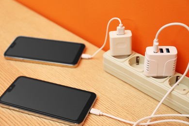 Photo of USB adapters with cables plugged into power strip charging smartphones on wooden table near orange wall, closeup