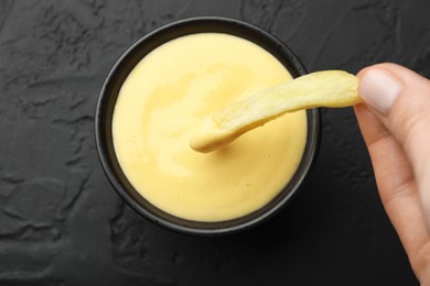 Photo of Woman dipping French fry into cheese sauce at black table, top view