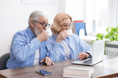 Photo of Senior man and mature woman drinking coffee while watching something on laptop at home. Happy couple