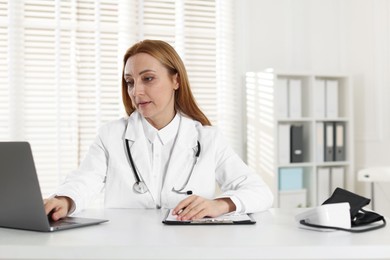 Photo of Cardiologist with stethoscope using laptop at white table in clinic