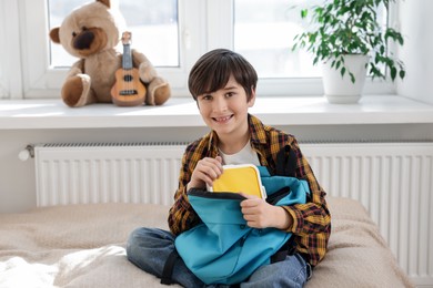 Photo of Boy packing lunch box into backpack on bed at home