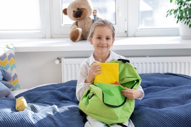 Photo of Girl packing book into backpack on bed indoors