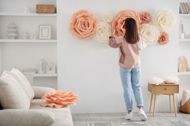 Photo of Woman decorating wall with beautiful paper flowers at home, back view