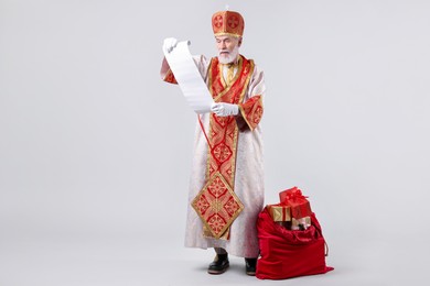 Photo of Saint Nicholas with wish list and sack full of gifts on light grey background