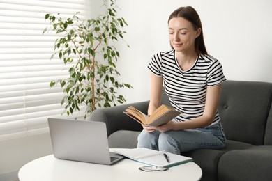 Photo of Woman with book and laptop learning at table on sofa indoors. Self-study