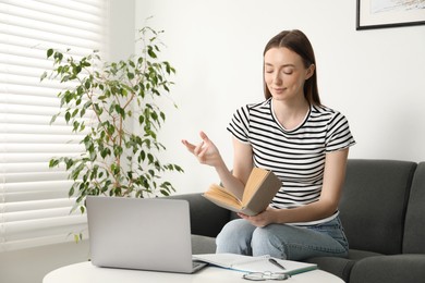 Photo of Woman with book and laptop learning at table on sofa indoors. Self-study