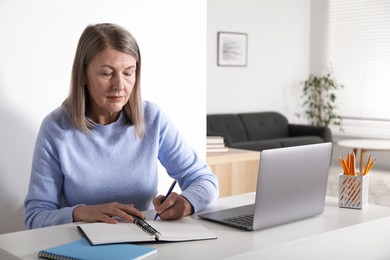 Photo of Woman learning online using laptop and taking notes at table indoors. Self-study