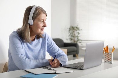 Photo of Smiling woman learning online using laptop and taking notes at table indoors. Self-study