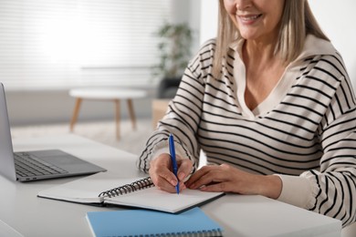 Photo of Smiling woman learning online using laptop and taking notes at table indoors, closeup. Self-study