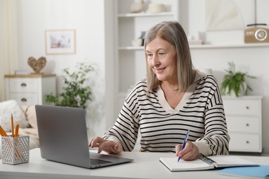 Photo of Smiling woman learning online using laptop and taking notes at table indoors. Self-study