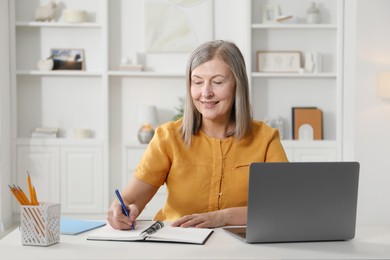 Photo of Smiling woman learning online using laptop and taking notes at table indoors. Self-study