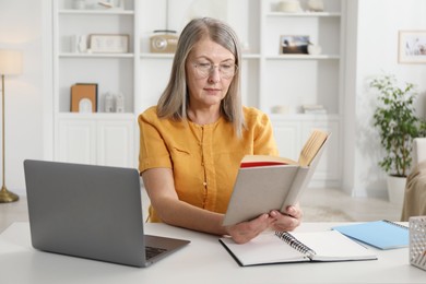 Photo of Woman with book learning near laptop at table indoors. Self-study