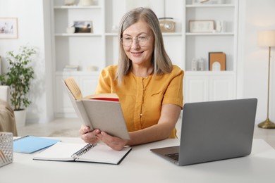 Photo of Woman with book learning near laptop at table indoors. Self-study