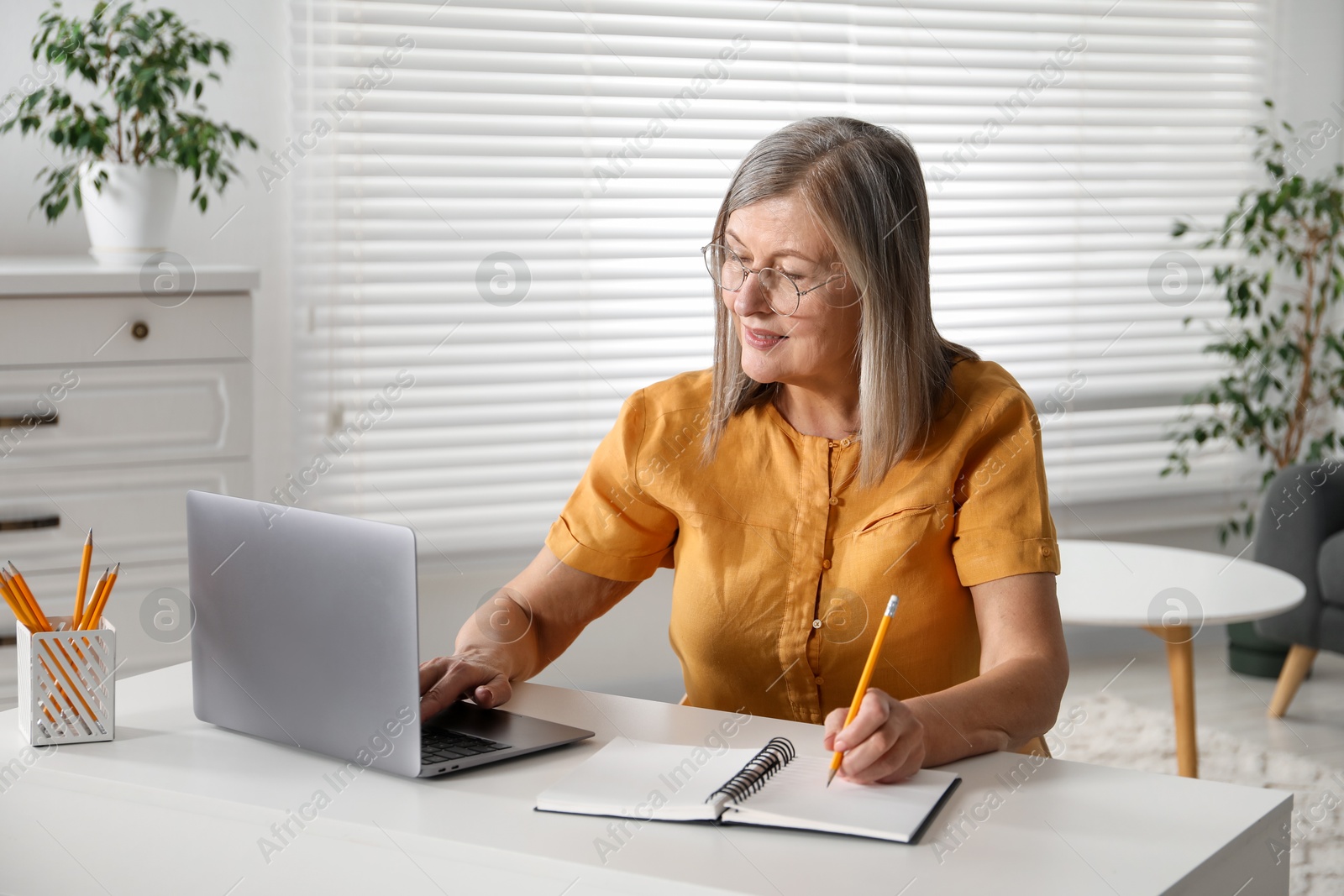 Photo of Smiling woman learning online using laptop and taking notes at table indoors. Self-study