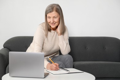 Photo of Smiling woman learning online using laptop and taking notes at table indoors. Self-study