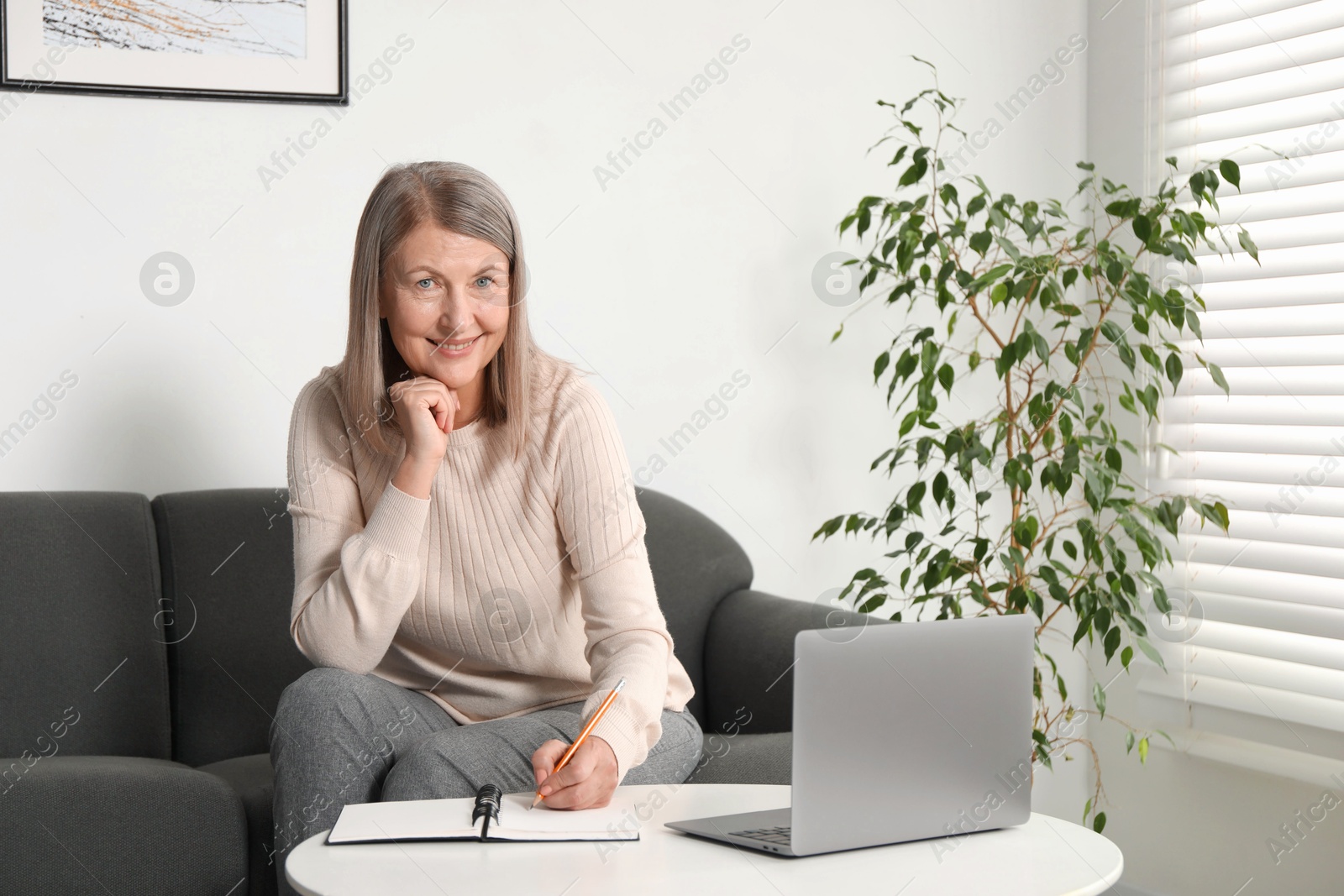 Photo of Smiling woman learning online using laptop and taking notes at table indoors. Self-study