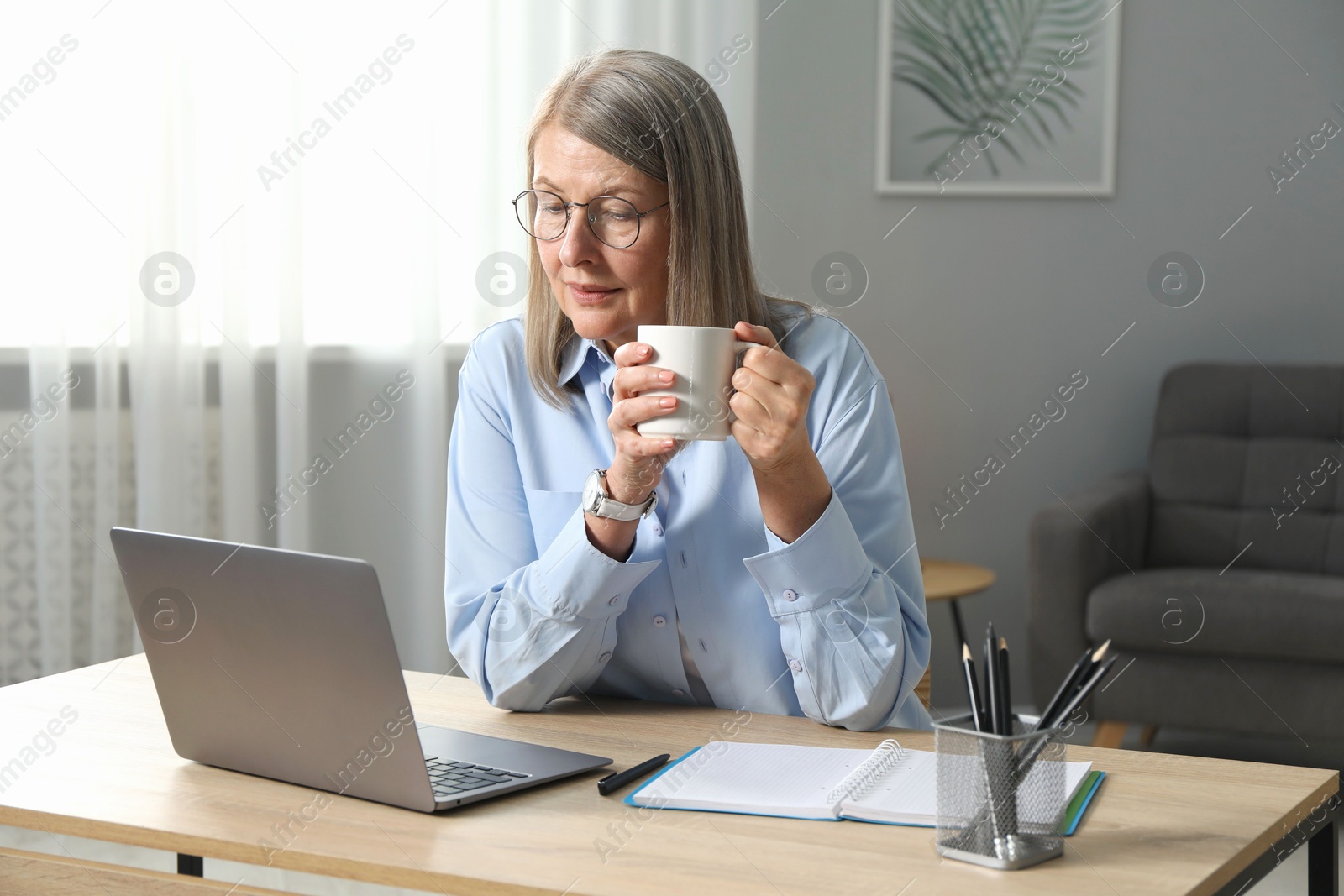 Photo of Woman with cup learning online using laptop at table indoors. Self-study