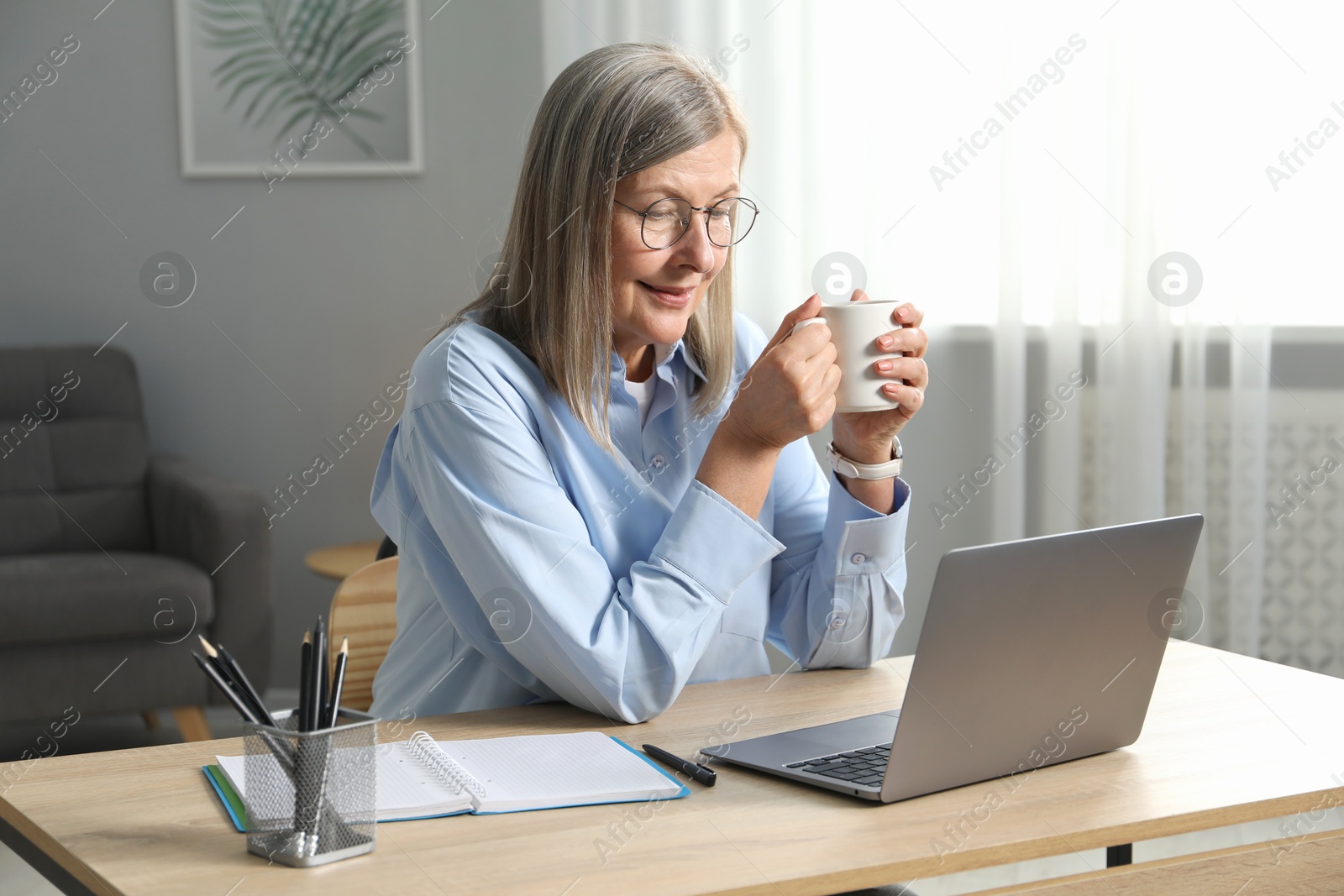 Photo of Woman with cup learning online using laptop at table indoors. Self-study