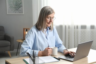 Photo of Woman with cup learning online using laptop at table indoors. Self-study