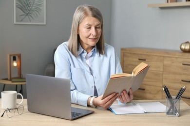 Photo of Woman with book learning near laptop at table indoors. Self-study