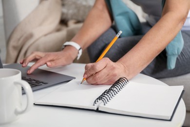 Photo of Woman learning online using laptop and taking notes at white table indoors, closeup. Self-study