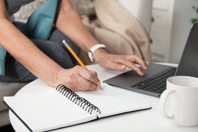 Photo of Woman learning online using laptop and taking notes at white table indoors, closeup. Self-study