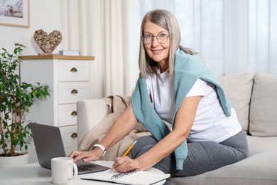 Photo of Smiling woman learning online using laptop and taking notes at table indoors. Self-study