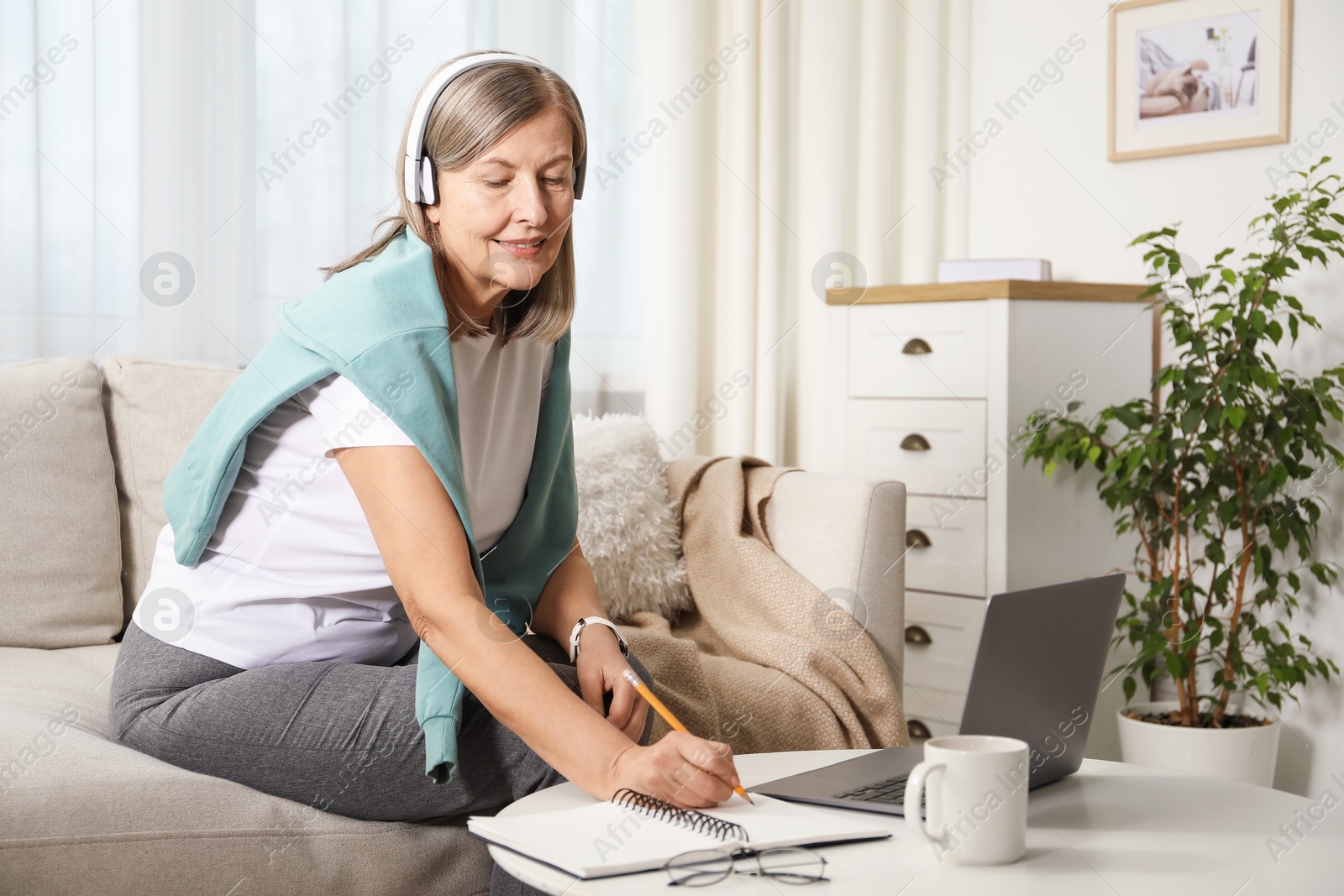 Photo of Smiling woman learning online using laptop and taking notes at table indoors. Self-study