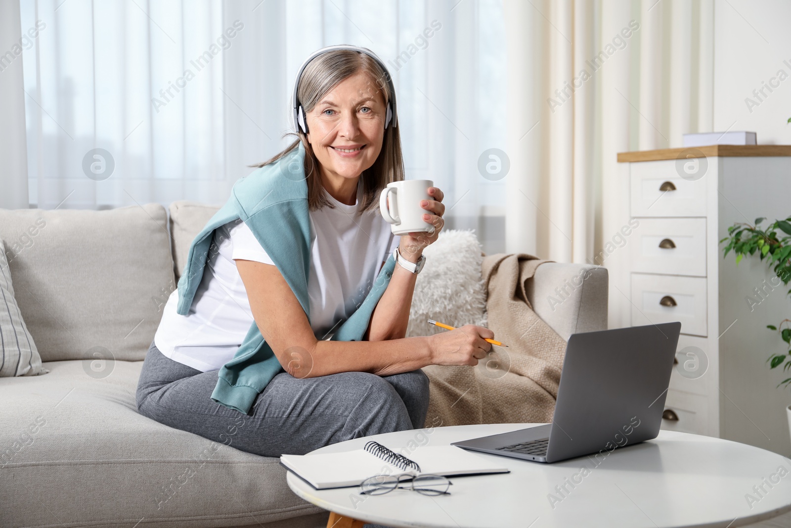 Photo of Smiling woman with cup learning online using laptop at table indoors. Self-study