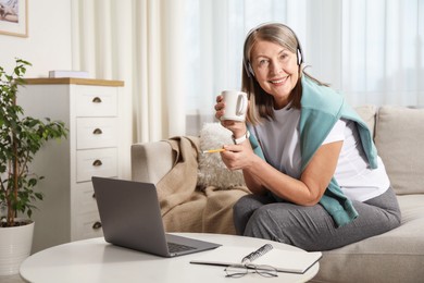 Photo of Smiling woman with cup learning online using laptop at table indoors. Self-study